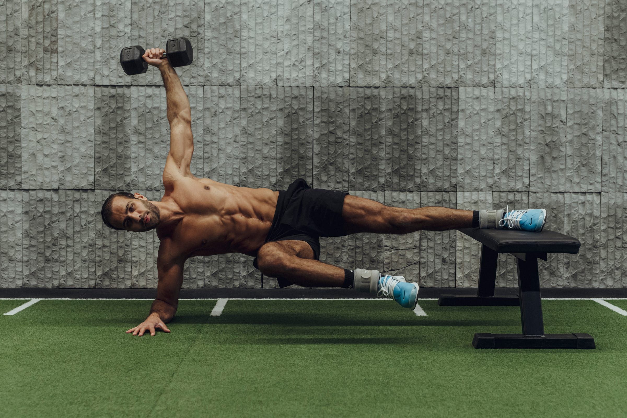 Sultan in a side plank on a bench, pressing a dumbbell overhead